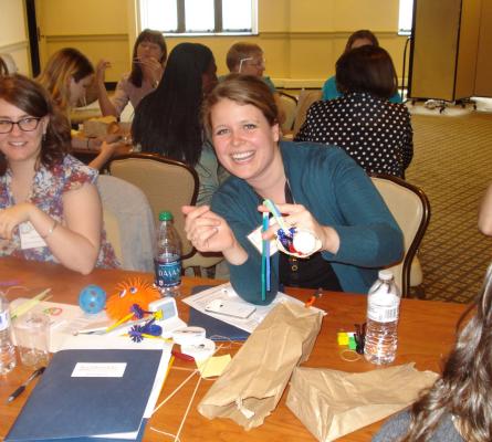 Photo of two women sitting at a table. One woman is holding something made of straws and pipe cleaners