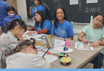 A group of adults and children sitting at a table. One adult is smiling at the camera, children are coloring