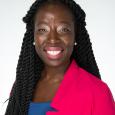 A Black woman with long dark braided hair wearing a pink blazer on top of a blue top