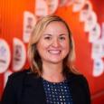 Anna Yudina Headshot - woman with blond hair, wearing blue jacket and blue shirt with polka dots standing in front of orange wall with white half circles