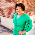 Stacy Brown - woman with short curly hair wearing a green dress standing in front of a brick wall