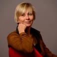 Claudine Schmuck - woman with short, blonde hair wearing brown jacket sitting on red chair