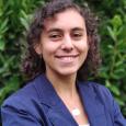 Vitoria Acerbi - Woman with short curly hair wearing blue jacket in front of green plants