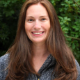 Alyssa Bar - woman with long brown hair swearing a gray sweater standing in front of a green hedge