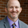 Joshua Sneideman - man with short hair and goatee wearing a purple shirt, multicolored tie, and gray suit jacket