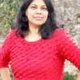 Dr. Lalitha Krishnamoorthy - woman with dark hair wearing red shirt standing in front of a stone wall