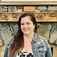 Lisa Broomall - woman with long brown hair wearing a jean jacket standing in front of a stone fireplace