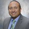 Snehal Bhakta - Headshot of man with black hair wearing a gray suit with green plaid tie