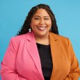 Woman with long dark hair in braids wearing pink and orange blazer in front of a blue background