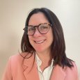 Yvette Mere-Cook - photo of woman with brown hair and glasses, wearing a white shirt and pink blazer