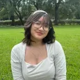 Teen girl with short dark hair and glasses sitting in a green park with trees